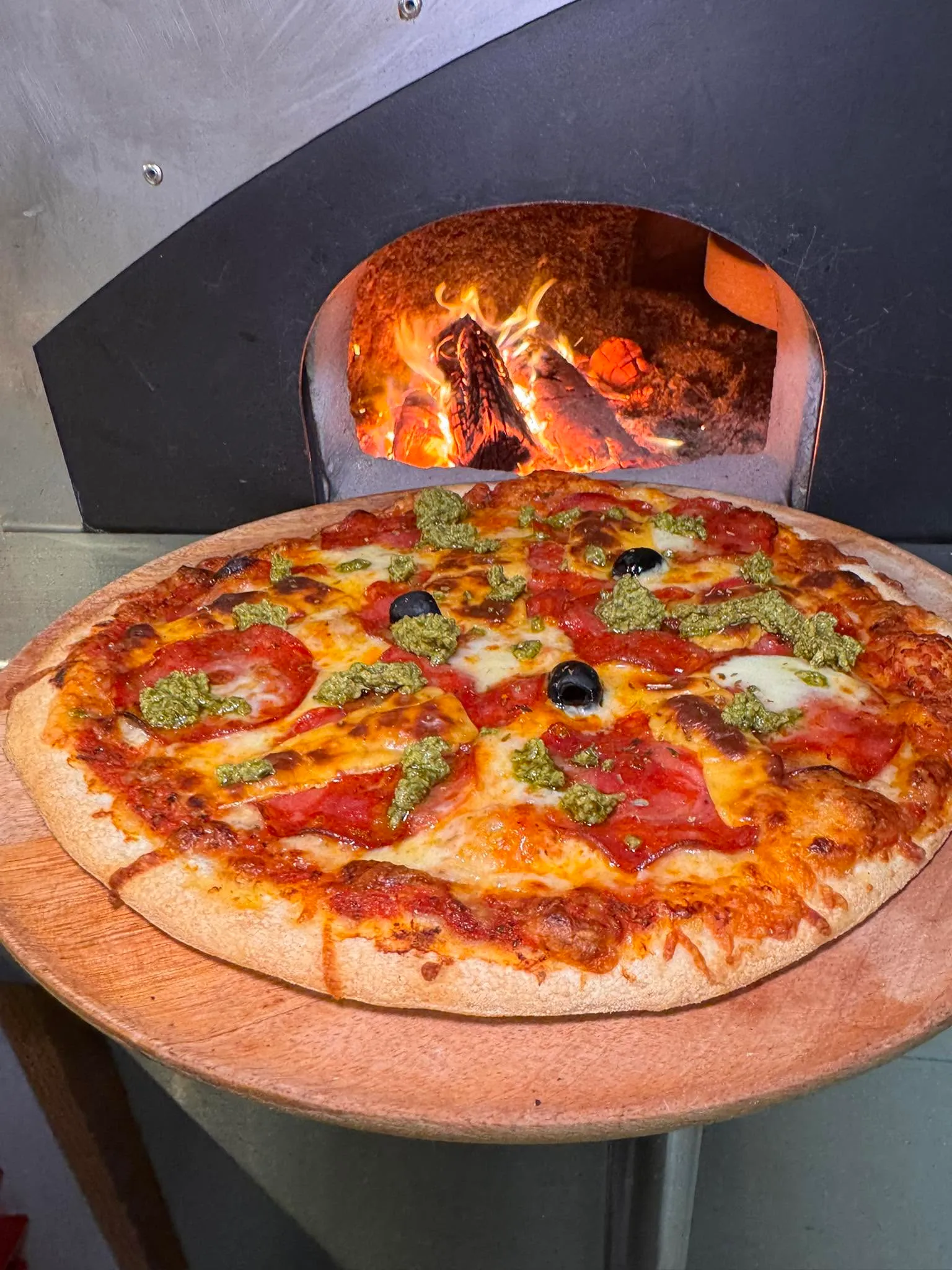 Pizza artisanale fraîchement sortie du four à bois, généreusement garnie de tranches de pepperoni, de touches de pesto au basilic, de fromage fondu et d'olives noires, présentée sur une pelle en bois.